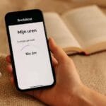 Hand met een telefoon waarop de BookBeat app de resterende luisteruren laat zien, met oortjes en een boek op de bank.