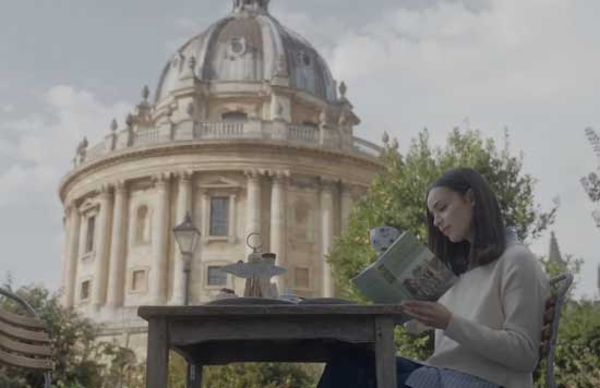 Een jonge vrouw leest een boek aan een tafel buiten in Oxford, met de Radcliffe Camera op de achtergrond, in een scene uit de romantische film My Oxford Year op Netflix.